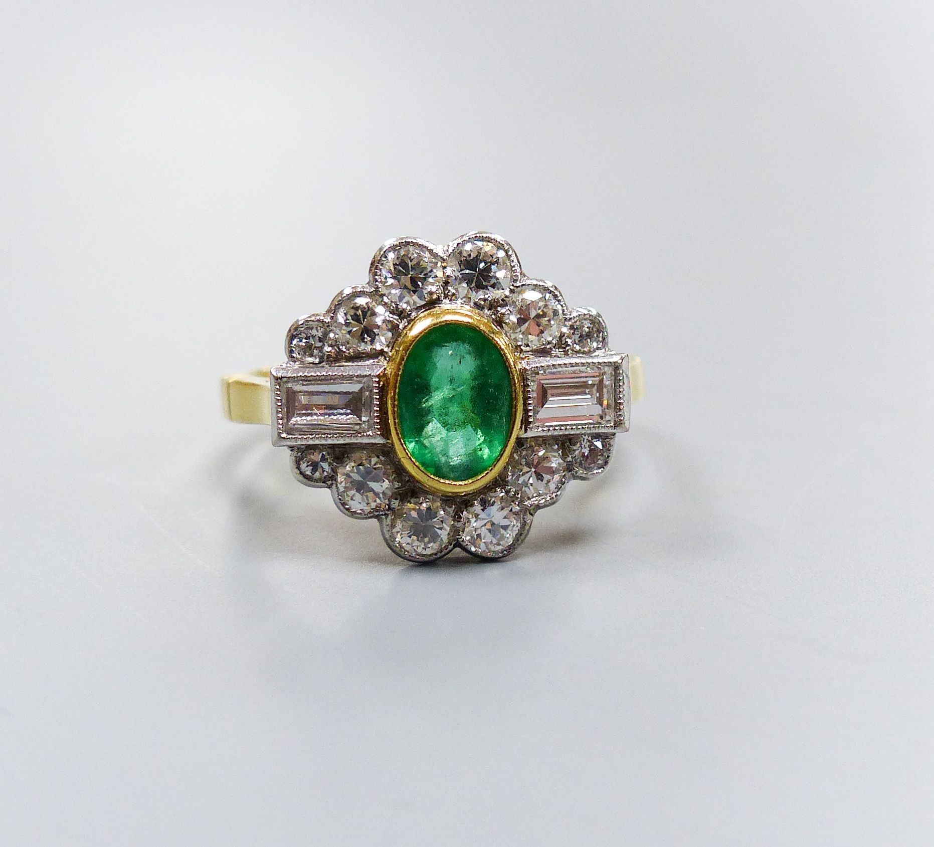 A modern 18ct gold, emerald, baguette and round cut diamond set shaped oval cluster ring, size L/M, gross weight 4.4 grams.